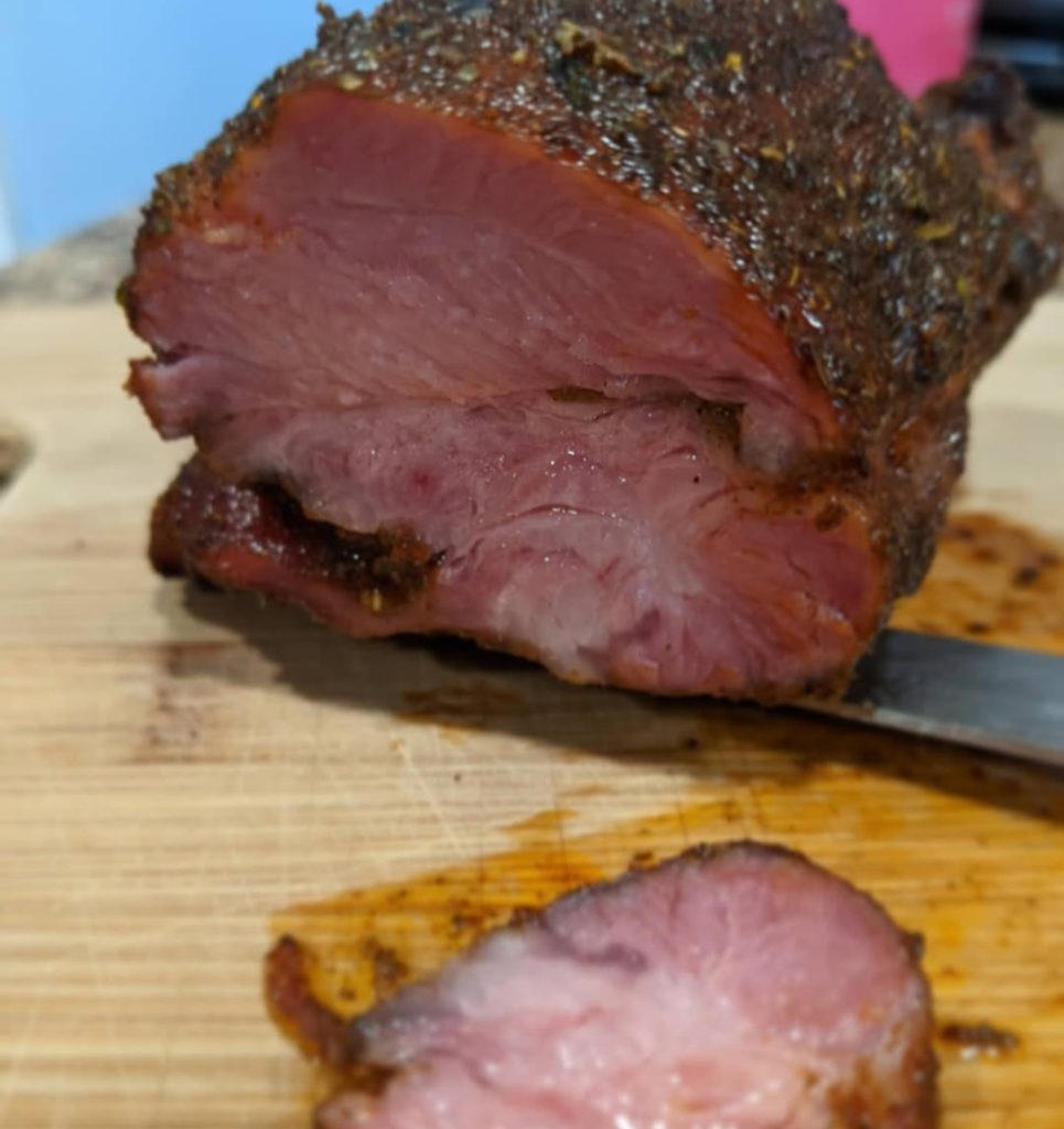 A close-up of sliced, smoked Tasso ham with a seasoned crust and tender, pink interior on a wooden cutting board.