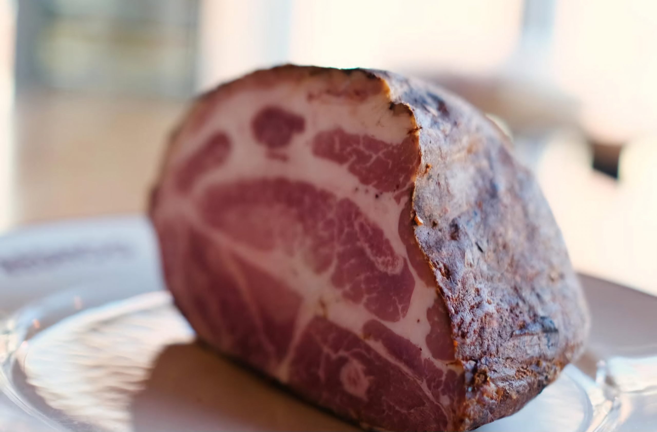 A close-up of a whole piece of Tasso ham, showcasing its marbled interior and smoky, seasoned exterior, resting on a white plate.