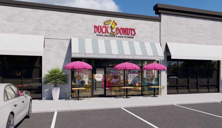 Duck Donuts storefront with a striped awning, logo, pink umbrellas, and outdoor seating.