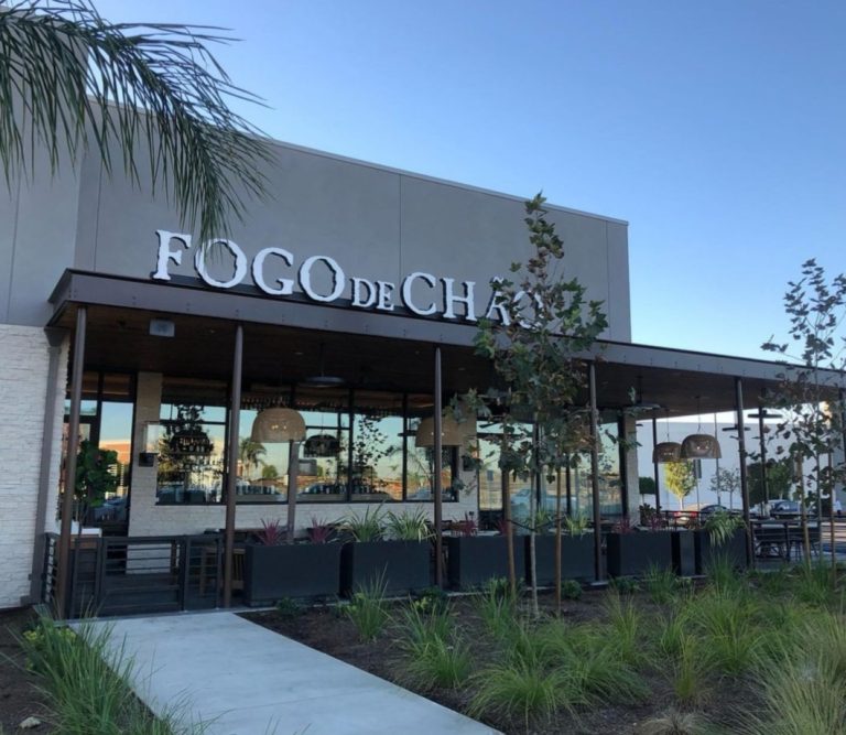 Exterior view of a Fogo de Chão restaurant with large windows, outdoor seating, and a prominent sign, surrounded by greenery under a blue sky.