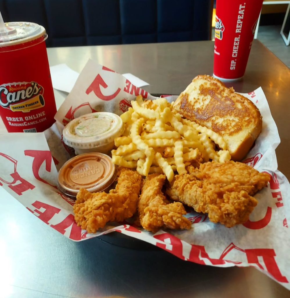 Raising Cane's menu with prices showcasing crispy chicken tenders, crinkle-cut fries, coleslaw, Texas toast, and signature dipping sauces, alongside a Raising Cane's drink cup.