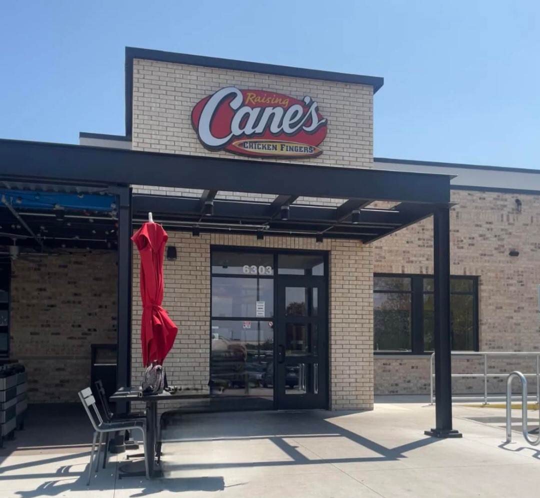 Exterior of a Raising Cane's Restaurant with a brick facade and shaded outdoor seating, showcasing the Raising Cane's sign above the entrance.