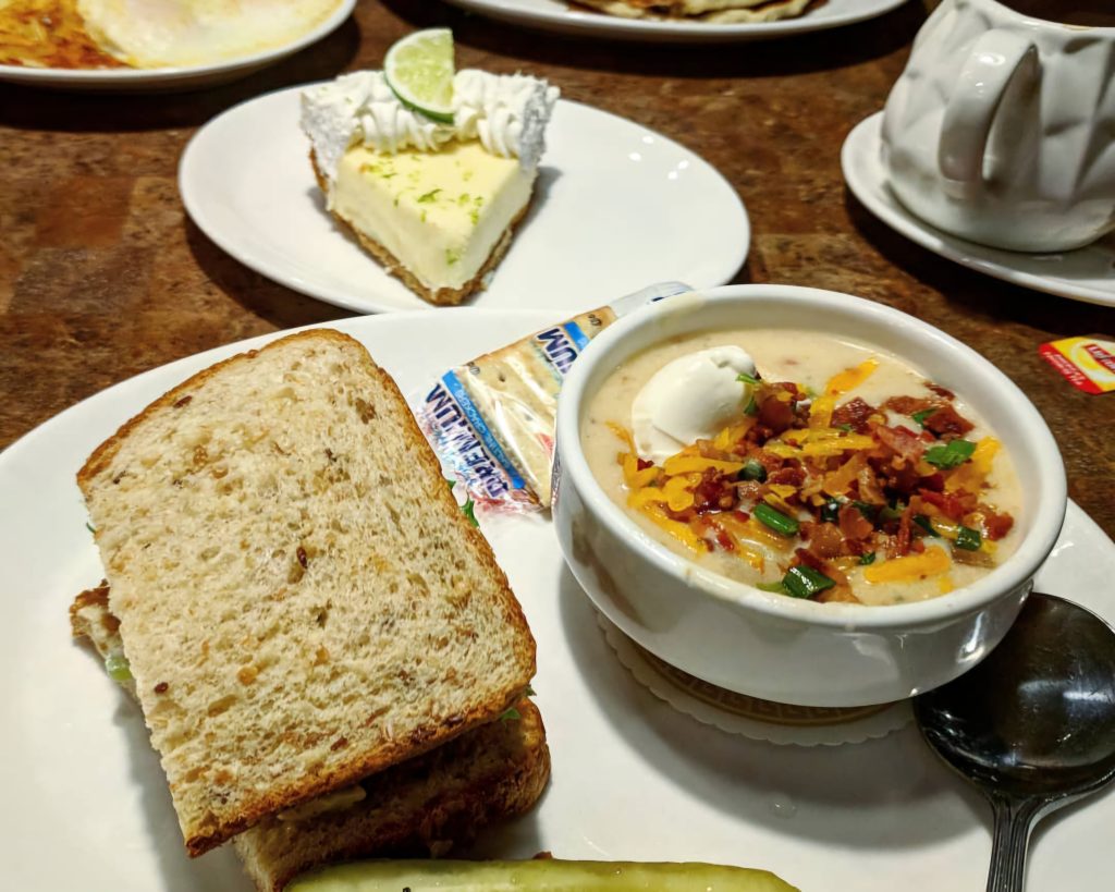 A delicious meal at Shari's Café & Pies, featuring a hearty sandwich, creamy soup topped with cheese and bacon, and a slice of key lime pie in the background.