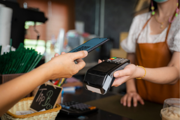 Customer using a smartphone for an Apple Pay payment at a store, with a tip box visible on the counter and a cashier holding the payment terminal.