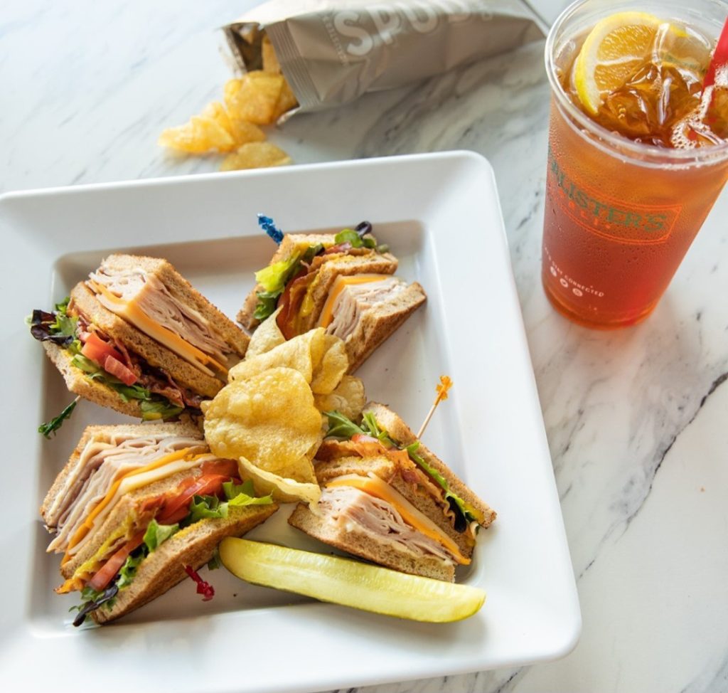 A McAlister’s Deli club sandwich platter served with crispy potato chips, a pickle spear, and a refreshing iced tea garnished with lemon, placed on a white plate on a marble table.